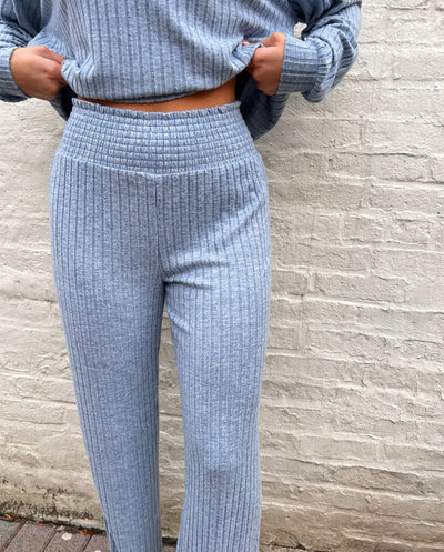 Person wearing a blue ribbed sweater and pants against a brick wall.